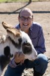 Ken Ramirez is kneeling down with his hand on the head of a donkey next to him. Ken is smiling at the camera.