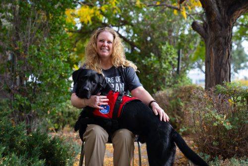 A woman is sitting in a chair outside. A black labrardor retriever has its front paws across her lap and its rear paws are on the ground.