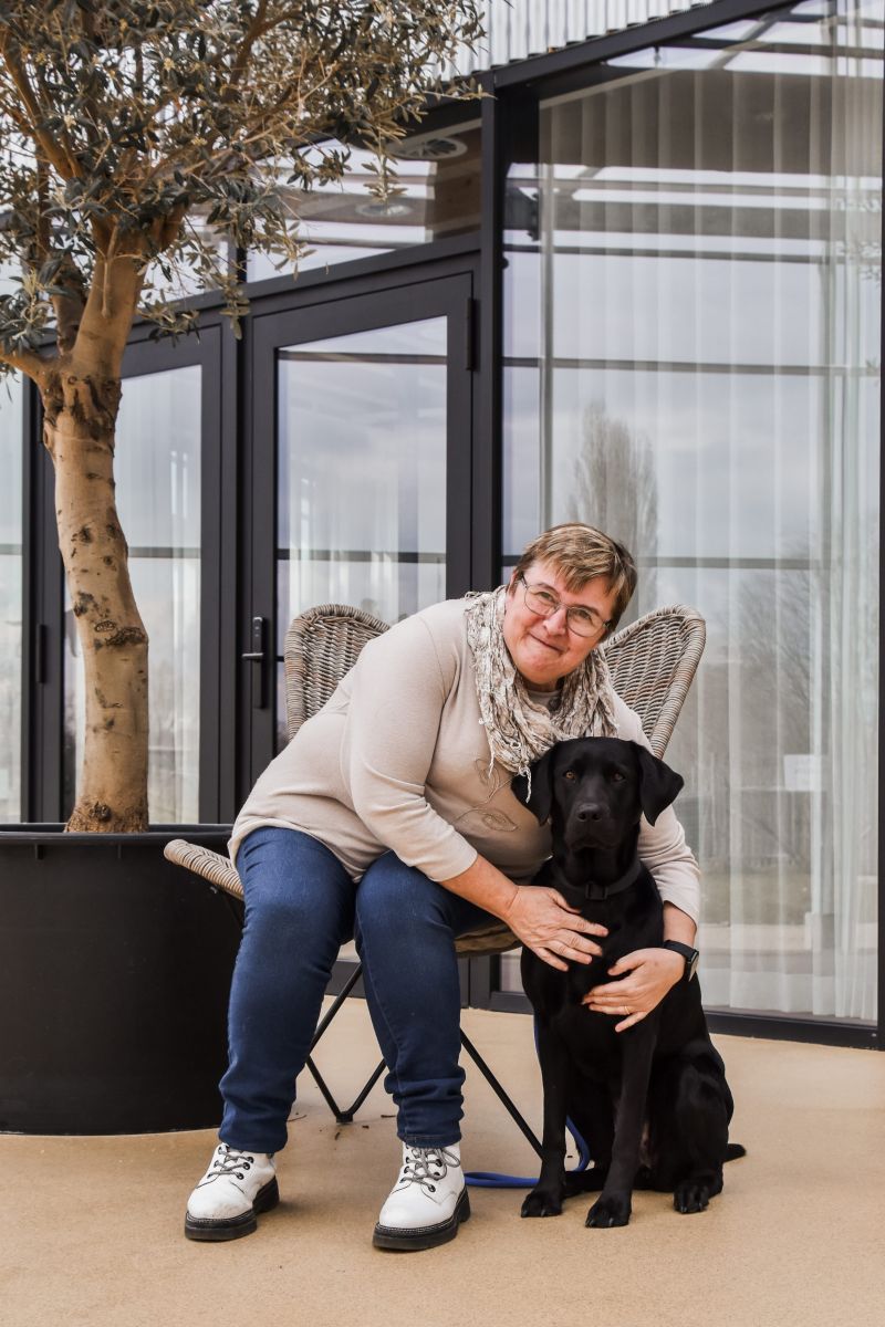 Reinhilde is sitting in a chair leaning over with her arms around Era, a black labrador sitting next to her chair.