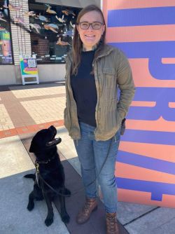 Haley is standing smiling at the camera. There is a black labrador sitting next to her looking up at her. 