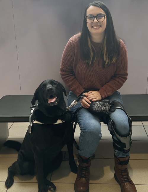 Kaitlyn sitting on a bench smiling at the camera. A black labrardor retriever is sitting next to her. 