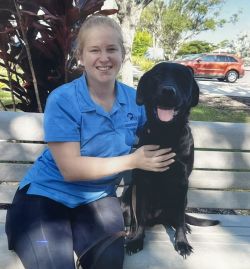 Katherine is sitting on a bench smiling at the camera. Her arms are around a black labrardor