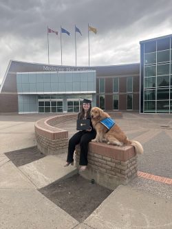 Kaylee is sitting outside Medicine Hat College holding a diploma. There is a golden retriever in assistance dog jacket with her