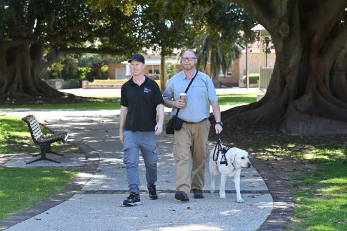 Kris is walking through a park. Wes is walking next to him. A OPK9 staff person is walking next to Kris.