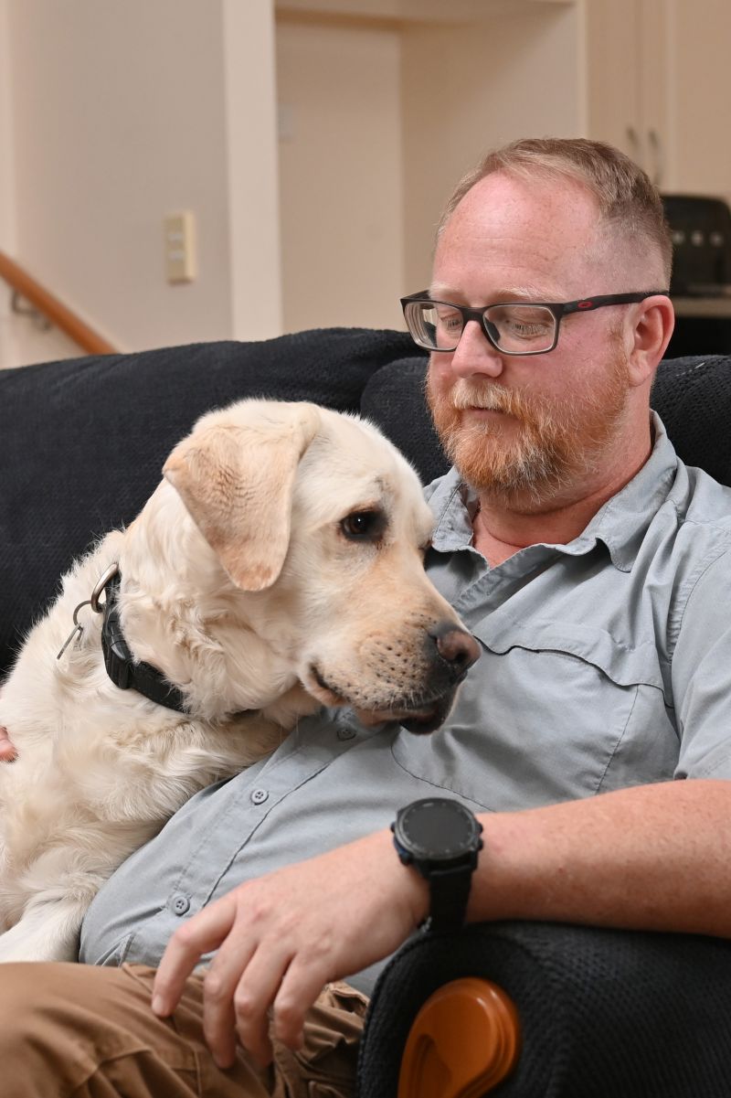 Kris is sitting on a couch looking down at his dog Wes who is sitting next to him.