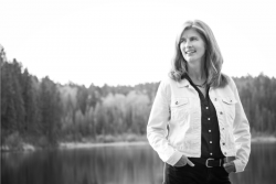A black and white photo of Kristina Stanley. There is a lake and trees behind her. 