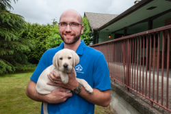 Matthias is smiling at the camera. He is holding a yellow labrador retriever puppy in his arms. Behind him is a house and yard.