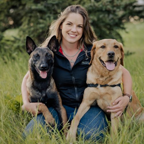 Miranda is sitting in a field smiling at the camera. She has a dog sitting on either side of her.