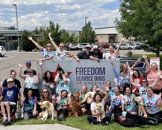 The staff, with multiple dogs, at Freedom Service Dogs of America is all gathered around the sign to their building. Most people are wearing a tie-dyed t-shirt.