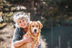 Sandra is bending over, arms around a golden retriever. Both are looking at the camera