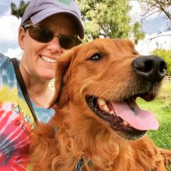Stephanie, in a hat and sunglass is smiling at the camera. A golden retriever with its mouth open is in front of her