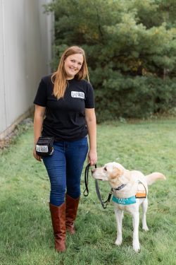 Sydney is standing outside, smiling at the camera. There is a yellow labrador retriever standing next to her in a service dog jacket.