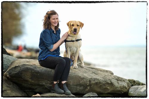 A woman is sitting on a rock next to a yellow labrador retriever. 