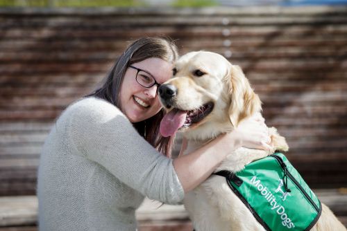 A woman is hugging a yellow retriever with green assistance dog jacket on.