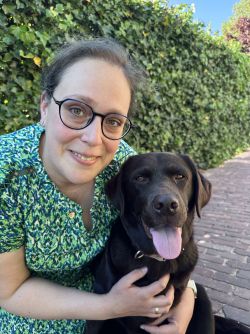 Corrien and her dog are posing for the camera. They are outside with a wall of ivy behind them. 