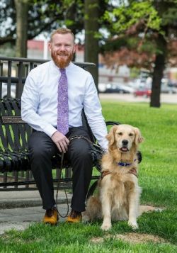 David is sitting on a bench outside. Next to him is a golden retriever in harness.
