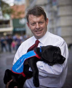 Richard is holding a black labrador puppy in his arms. The puppy has an assistance dog jacket on. Richard is smiling at the camera.