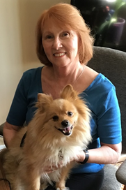 Sheila O'Brien is sitting on a chair smiling at a camera. On her lap is a pomeranian dog. 