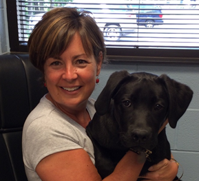 Jenny Barlos is smiling at the camera. She is holding a young black labrador retriever in her lap. 