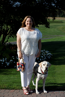 Valerie Cramer is smiling at the camera. Next to her is a yellow labrador retriever in an assistance dog jacket. 