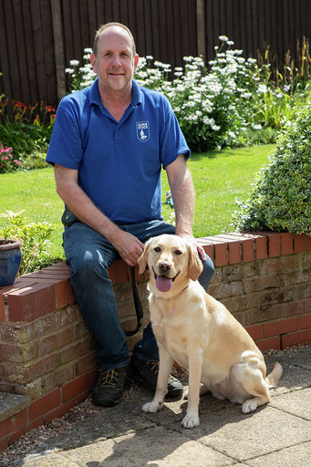 David Bailey is sitting on a short brick wall smiling at the camera. He has a yellow labrador retriever sitting in front of him.
