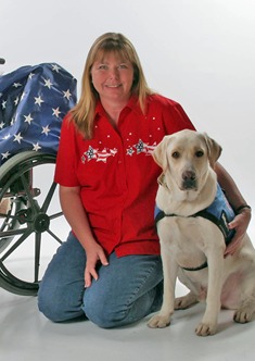 Lori Stevens is kneeling on the ground and smiling at the camera. She has her arm around a yellow labrador retriever that has a blue jacket on. There is a wheel chair on the other side of Lori's body. 