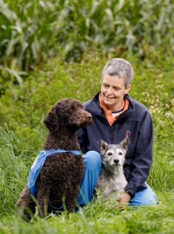 Elisabeth is kneeling on the ground smiling at the two dogs sitting by her.