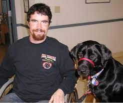 A man in a wheelchair is next to his service dog, a black labrador retriever. 