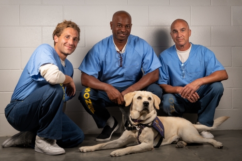 Three inmate handler puppy raisers are kneeling behind a yellow Labrador Retriever. The men are smiling at the camera. The dog is wearing a purple assistance dog vest and is lying down. 
