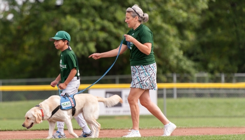 A young boy is holding a handle attached to a blue assistance dog vest on a yellow Labrador Retriever. An adult woman is walking behind the boy and dog holding the dog's leash. The team is on a baseball field and the people are wearing uniform shirts. 