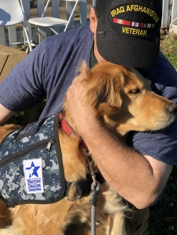 A man wearing a black hat that has "Iraq Afghanistan Veteran" printed on it is hugging a golden retriever. The dog is wearing a camouflage pattern assistance dog vest.  