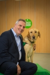 Peter Gorbing is sitting with his arm around a yellow Labrador retriever. The dog is wearing a green assistance dog vest.